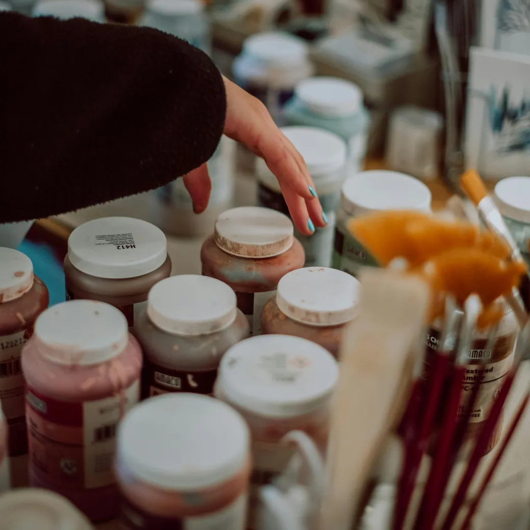 A hand reaching across a table crowded with jars of coloured paint and glaze, brushes and mixing sticks
