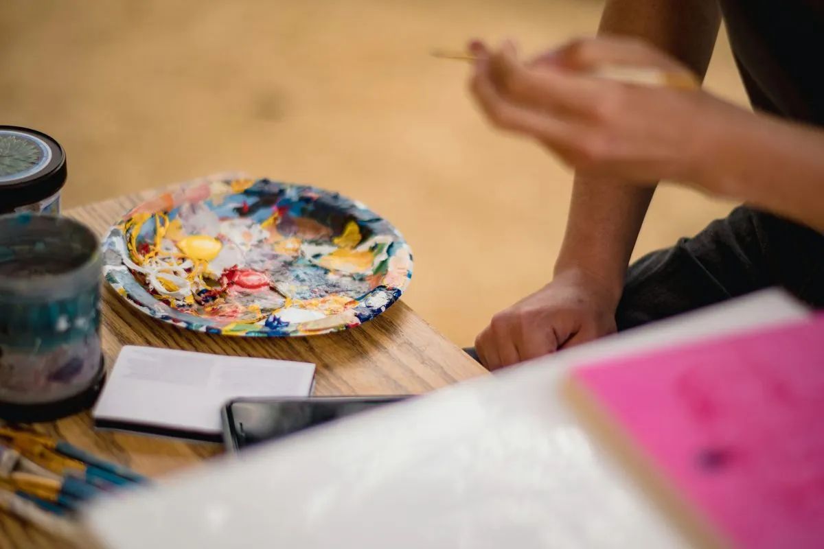 A paint-covered palette, brushes and a notebook on a studio table, with a participant painting just beyond